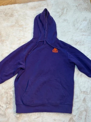 Lila hoodie från Kappa - Säljer en lila hoodie från Kappa med klassisk huva och magficka. Kappa-loggan i orange brodyr på bröstet ger en sportig vibe. Tillverkad i mjukt bomullsmaterial som är skönt att ha på sig. Perfekt för dig som gillar streetwear och vill ha något bekvämt!