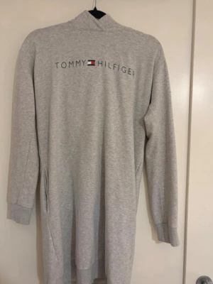 Grå lång hoodie från Tommy Hilfiger! - Mysig grå lång hoodie från Tommy Hilfiger med logga och flagga på ryggen. Tröjan har huva, långa ärmar och ribbade muddar. Perfekt för chill dagar hemma eller som loungewear. Tillverkad i mjukt sweatshirtmaterial!