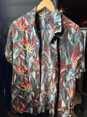 Blommig kortärmad skjorta från H&M - Säljer en färgstark kortärmad skjorta från H&M med blommigt mönster i rött, gult och grönt på brun botten. Skjortan har klassisk krage, knappar framtill och är gjord i ett lätt bomullsmaterial. Perfekt för dig som vill sticka ut lite extra!