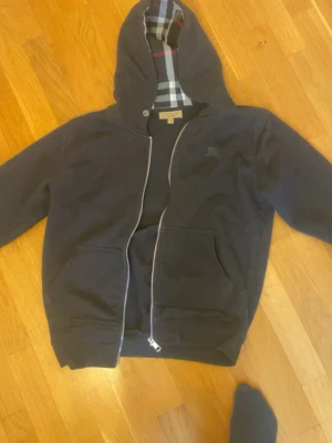 Svart hoodie från Burberry med dragkedja - Snygg svart hoodie från Burberry med klassiskt rutigt foder i huvan och dragkedja framtill. Hoodien har två fickor och diskret Burberry-logga på bröstet. Perfekt för en stilren och avslappnad look. Tillverkad i mjukt bomullsmaterial.