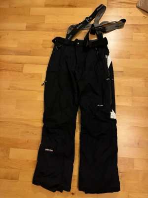 Täckbyxor - Svarta täckbyxor med justerbara hängslen som också är avtagbara. Många bra fickor med dragkedja. Perfekta för vinterns alla äventyr i backen. Strl S.