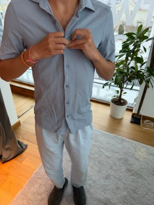 Ljusblå kortärmad skorta - Säljer en ljusblå kortärmad skjorta med klassisk krage och knappar framtill. Skjortan har en avslappnad passform och är tillverkad i ett mjukt, tunt material som känns skönt mot huden. 