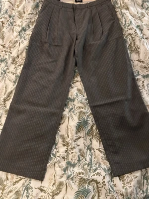 Grå kritstrecksrandiga kostymbyxor - Säljer ett par grå kritstrecksrandiga kostymbyxor från Jack & Jones. Byxorna har bred passform, raka ben och bälteshällor. Klassisk look med två bakfickor med knapp och diskret dragkedja framtill. Perfekta för en clean och stilren outfit. Aldrig använda!