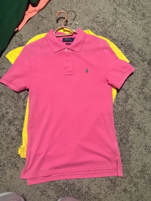 Rosa pikétröja från Polo Ralph Lauren - Säljer en klassisk rosa pikétröja från Polo Ralph Lauren med liten grön logga broderad på bröstet. Tröjan har krage, korta ärmar och knappar vid halsen. Perfekt till sommaren för en färgglad och sportig look.