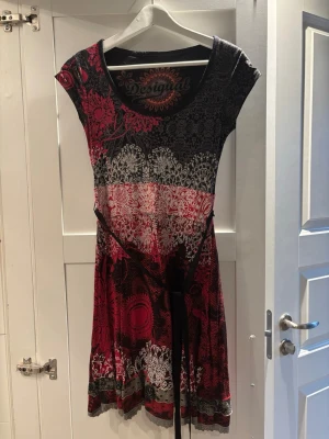 Desigual mönstrad kortklänning - Säljer en färgstark kortklänning från Desigual med coolt blommigt och mandalainspirerat mönster i svart, rött och grått. Klänningen har v-ringad hals, korta ärmar och ett knytband i midjan som ger en snygg siluett. Perfekt för dig som gillar unika prints!