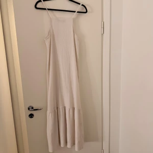Lång vit klänning med smala axelband - Säljer en lång vit klänning med smala axelband och lätt rynkad struktur i tyget. Klänningen har en rak passform upptill och ett volangparti nedtill som ger extra rörelse. Materialet känns mjukt och luftigt, perfekt för varma dagar.