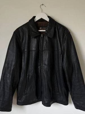 Svart skinnjacka från Columbia XL - Svart skinnjacka från Columbia i klassisk modell med krage och dragkedja framtill. Jackan har två sidofickor och brunt innerfoder. Perfekt för dig som gillar en stilren och tidlös look. Passar dig som vill ha en jacka med lite loose fit. 7Y9