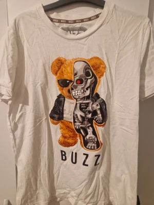 Vit t-shirt med robotnalle från Buzz - Vit t-shirt från Buzz Sneaker Station i storlek XS med en stor, färgglad print av en nalle som är halvt robot och har solglasögon. Rund hals och korta ärmar. Materialet känns mjukt och är troligtvis bomull. Perfekt för dig som gillar coola och unika motiv.