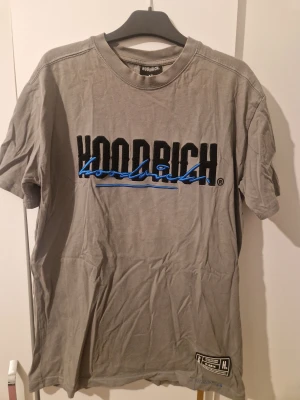 Grå Hoodrich t-shirt med blå detaljer - Säljer en grå t-shirt från Hoodrich med fet broderad logga i svart och blått på bröstet. T-shirten har korta ärmar, rund hals och cool streetwear-patch längst ner. Materialet är mjuk bomull och det finns blå sömmar vid nederkanten för extra stil.