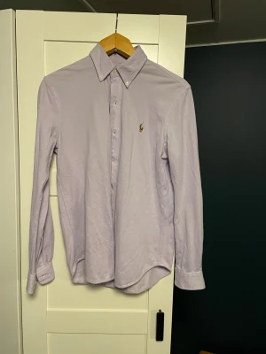 Ljuslila skjorta från Ralph Lauren - Säljer en stilren ljuslila skjorta från Ralph Lauren, modell Knit Oxford. Skjortan har klassisk krage med knappar, lång ärm och det ikoniska broderade logot på bröstet. Materialet är mjukt och känns som bomull, perfekt för en clean look.