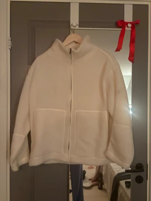 Vit teddyjacka från Stories S36/38 - Mysig vit teddyjacka från Stories i storlek S36/38. Jackan har hög krage, två stora fickor framtill och dragkedja hela vägen. Perfekt för kalla dagar och har en loose passform som ger en chill vibe.