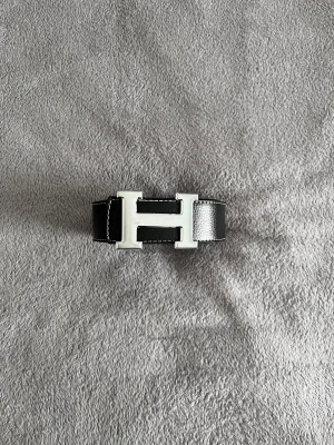 Hermès bälte  - Säljer ett stilrent svart skärp från Hermès med ett stort H-format spänne i silverfärgad metall. Skärpet är gjort av äkta skinn med tydliga vita sömmar längs kanterna och har en klassisk, rak form. Perfekt för att lyfta din outfit med en lyxig detalj.