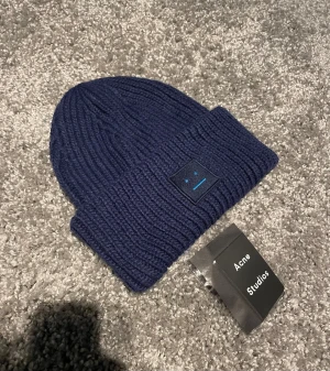 Marinblå stickad mössa Acne Studios - Säljer en marinblå stickad mössa från Acne Studios med det klassiska ansikts-emblemet framtill. Mössan är ribbstickad och har en bred uppvikt kant. Perfekt för att hålla värmen och ge din outfit en clean vibe.