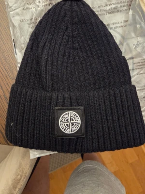 Svart ribbad mössa från Stone Island - Säljer en svart ribbstickad mössa från Stone Island med klassisk logopatch framtill. Mössan är mjuk och har en uppvikt kant för extra stilpoäng. Perfekt för dig som vill ha en clean och snygg look under kalla dagar.