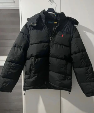 Svart pufferjacka från Polo Ralph Lauren - Svart pufferjacka från Polo Ralph Lauren med röd broderad logga på bröstet. Jackan har huva, två sidofickor med dragkedja, perfekt för kalla dagar.