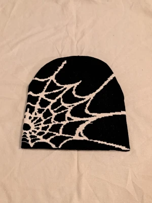 Svart stickad mössa med spindelnätsmönster - Säljer en svart stickad mössa med ett vitt spindelnätsmönster som täcker ena sidan. Mössan är mjuk och stretchig, perfekt för dig som gillar lite edgy stil. Passar dig som vill sticka ut och gillar coola detaljer.