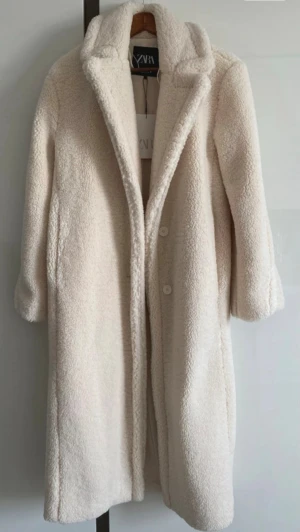 Teddyjacka från Zara - Säljer en lång, beige/cremefärgad teddyjacka från Zara i storlek XS. Jackan har krage, knappar framtill och är gjord i mjukt fleece-material som ger en fluffig och mysig känsla. Helt ny och säljs inte längre i Zara affären.