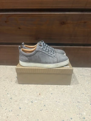 Grå mocka sneakers från Louboutin - Snygga grå sneakers från Christian Louboutin i mjuk mocka med klassisk vit platt sula och ikonisk röd undersida. Skorna har rund tå, snörning och beige innersida. Perfekta för dig som vill ha en clean och lyxig look med street vibe. Allt på bilderna skickas med! Priset är såklart alltid diskuterbart🤗