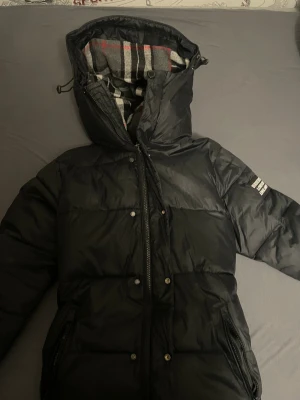 Svart pufferjacka från Burberry - Svart pufferjacka från Burberry med huva och dragkedja framtill. Insidan har klassiskt rutigt Burberry-foder i grått, vitt och rött. Jackan har tryckknappar med Burberry England-text och en patch på ärmen. Perfekt för kalla dagar.