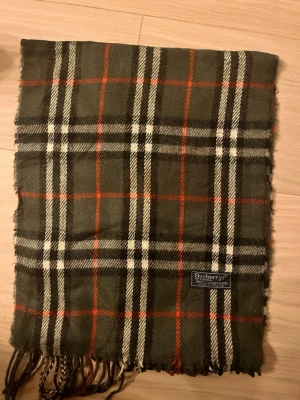 Rutig ullväst från Burberrys - Snygg rutig väst från Burberrys i mörkgrön och röd färg med inslag av vitt och svart. Västen är tillverkad i 100% lammull och har klassiskt rutmönster. Perfekt för dig som gillar tidlös design och vill ha något som sticker ut lite extra.