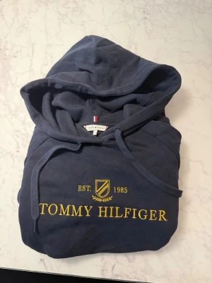 Mörkblå Tommy Hilfiger Hoodie - Säljer denna schyssta Tommy Hilfiger hoodien som aldrig kommit till användning. Storlek S fast den är rätt liten i storlek så skulle säga XS