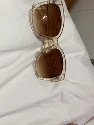 Ingen väst på bilden - Bilden visar ett par solglasögon med transparenta bågar och bruna glas. Ingen väst finns på bilden.