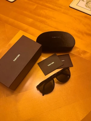 Tom Ford solglasögon - Säljer ett par snygga svarta solglasögon från Tom Ford. Modellen har klassisk form med breda bågar och mörka linser. Medföljer originalfodral, putsduk och box & kvitto. Perfekt accessoar för att höja din stil. 