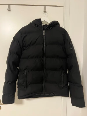 Svart pufferjacka Supply & Demand - Svart pufferjacka från Supply & Demand med huva och snygga detaljer som patch på ärmen. Jackan har dragkedja framtill och två fickor med dragkedja. Perfekt för kalla dagar och har en modern, loose passform.