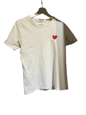 Comme des Garçons Play - Säljer en vit t-shirt från Comme des Garçons Play med det ikoniska röda hjärtat med ögon broderat på bröstet. T-shirten har rund hals, korta ärmar och är gjord i mjuk bomull. Perfekt för dig som gillar stilrena plagg🥂🍾🍾👍