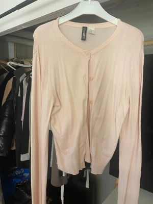 Ljusrosa kofta från Divided - Säljer en söt ljusrosa kofta från Divided med rund hals och långa ärmar. Koftan har knappar framtill och är gjord i mjukt, tunt material som känns skönt mot huden. Perfekt att slänga på över en topp när det är lite kyligt ute.