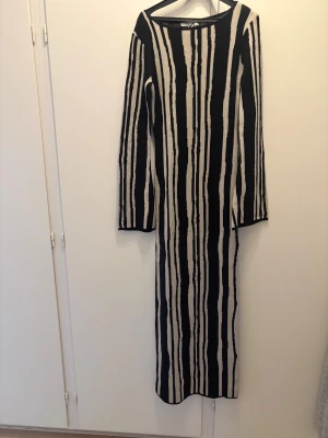 Randig långklänning från NA-KD - Snygg långklänning från NA-KD med svart och beige vertikalt randigt mönster. Klänningen har långärmade och är stickad i ett mjukt material som ger en skön känsla.