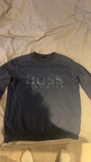 Blå slim fit tröja från Hugo Boss - Snygg blå tröja från Hugo Boss i slim fit-modell med stort BOSS-tryck på bröstet och som är i storlek S. Tröjan har rund hals, långa ärmar och ribbade muddar vid ärmslut och nederkant. Perfekt för dig som gillar stilrena och klassiska plagg med en modern touch.