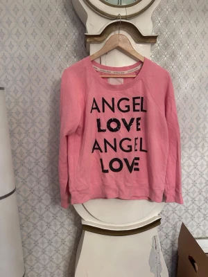 Rosa sweatshirt från Victoria's Secret - Mysig rosa sweatshirt från Victoria's Secret med svart text 'ANGEL LOVE' framtill. Tröjan har rund halsringning, långa ärmar och ribbade muddar. Perfekt för en avslappnad och trendig look.
