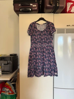 Odd Molly blå kortklänning med mönster - Supersöt kortklänning från Odd Molly i mörkblått med rosa paisley-mönster. Klänningen har puffiga kortärmar, rund halsringning med snörning och volangdetaljer nedtill. Tillverkad i mjuk bomull som känns skön mot huden. Från märket Odd Molly. Storlek 2 som enligt deras storleksguide motsvarar en M. Fin men den ör tyvärr något tvätt påverkad. 