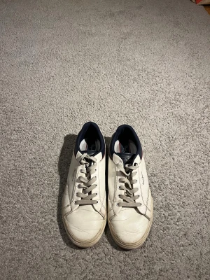 Vita sneakers från Pepe Jeans - Snygga vita sneakers från Pepe Jeans med marinblå detaljer och röd linje på sidan. Skorna har snörning, rund tå och platt sula. Ovandelen är i skinn och mocka, med mjuk textil på insidan. Perfekta för dig som gillar klassisk stil med en twist.