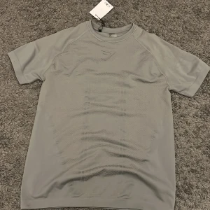 Grå Gymshark t-shirt onyx  - Snygg grå t-shirt från Gymshark med diskret logga på bröstet. Tillverkad i mjuk och lätt polyesterblandning som andas, perfekt för träning. T-shirten har korta ärmar och en klassisk rund hals. Enkel design med sportig vibe.