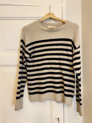 Randig stickad tröja från Vila - Säljer en snygg stickad tröja från Vila i storlek S. Tröjan är beige med svarta horisontella ränder, har rund hals och långa ärmar. Den är ribbstickad vid hals, ärmslut och nederkant, och har en normal passform. Perfekt för dig som gillar klassisk stil med en twist.