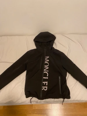 Svart Moncler vindjacka med huva - Svart vindjacka från Moncler med stor logga längs dragkedjan. Jackan har huva med snörning, två sidofickor med dragkedja och justerbar nederkant. Materialet är syntet och jackan har en clean, sportig look som passar perfekt till streetwear. Det är storlek M men passar mer som en S. Pris kan diskuteras!