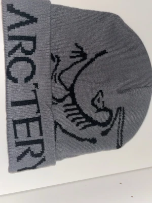Grå mössa från Arc'teryx - Säljer en snygg grå mössa från Arc'teryx med svart logga och grafiskt mönster framtill. Mössan är stickad i mjukt material och har en bred uppvikt kant med Arc'teryx-text. Perfekt för kalla dagar och outdoor-häng.