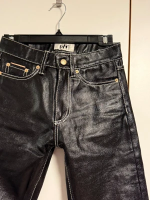 Straight skinnbyxor från Eytys - Säljer ett par svarta skinnbyxor från Eytys med vita sömmar, modell Cypress. Storlek 26/32. De har en straight passform, kontrasterande vita sömmar och guldfärgade detaljer vid fickorna. Materialet är jeans med en glansig finish som ger en cool look. Dragkedja och knappstängning framtill. 