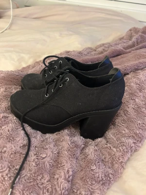 Svarta klackskor från Divided - Säljer ett par svarta klackskor från Divided med snörning och rund tå. Skorna har en rejäl plattform och är tillverkade i textil med syntetdetaljer vid hälen. Perfekta för dig som gillar chunky stil och vill ha lite extra höjd.