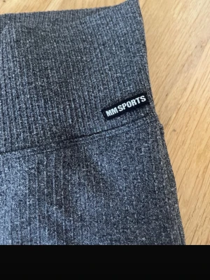 Grå ribbade leggings från MM Sports - Säljer ett par grå leggings från MM Sports i storlek L/XL. De är ribbade med bred, hög midja och har en mjuk, stretchig känsla som passar perfekt för träning. Materialet är syntet och designen är enkel och sportig.