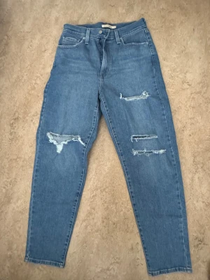 Levi's blå high waisted mom jeans - Säljer ett par blå high waisted mom jeans från Levi's med slitna detaljer och klassisk femficksdesign. Jeansen har raka ben, dragkedja och knapp framtill samt Levi's patch bak på midjan. Perfekt för dig som gillar en avslappnad och trendig look.