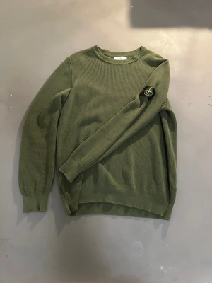 Grön stickad tröja från Stone Island - Snygg grön stickad tröja från Stone Island med klassisk rund hals och ribbade muddar. Tröjan har lång ärm och den ikoniska Stone Island-loggan på vänster ärm. Perfekt för dig som gillar streetwear och vill ha något stilrent och bekvämt.