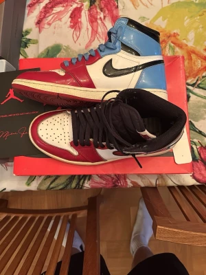 Nike Air Jordan 1 Retro High OG Fearless - Nike Air Jordan 1 Retro High OG Fearless i färgerna röd, vit, svart och ljusblå. Skorna har högt skaft, svart swoosh och snörning. Ovandelen är i glansigt skinn och designen är riktigt ikonisk med mixade färgblock. Perfekt för dig som vill sticka ut med din sneakerstil.  Går för runt 3500 upp till 7000 nya.