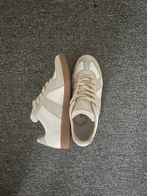 Beige och vita sneakers med mockadetaljer - Säljer ett par stilrena sneakers från Birkenstock i beige och vitt med snygga mockadetaljer och bruna gummisulor. Skorna har rund tå, snörning och en platt sula som gör dem riktigt sköna att gå i. Perfekta för dig som gillar minimalistisk och clean stil.