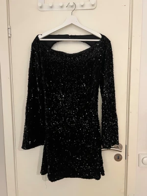 Svart glittrig paljettklänning - Säljer en svart kort klänning med långa ärmar, täckt av glittriga paljetter. Klänningen har en öppen rygg med snörning och en snygg, figurnära passform. Perfekt för dig som vill sticka ut med en glamorös look. Materialet är troligtvis polyester.
