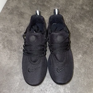 Nike Presto svarta sneakers - Svarta Nike Presto sneakers med mesh-ovandel och snygga plastdetaljer på sidorna. Skorna har snörning, rund tå och platt sula. Diskret Nike-logga på tån och bakre flärp för enkel påtagning. Perfekt för dig som gillar stilrena och sportiga sneakers.