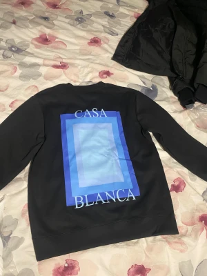 Svart sweatshirt från Casablanca - Svart sweatshirt från Casablanca med stort blått grafiskt tryck och texten 'Casa Blanca' på ryggen samt mindre tryck på bröstet. Tröjan har rund hals och långa ärmar, perfekt för dig som gillar stilrena och moderna plagg med en twist.