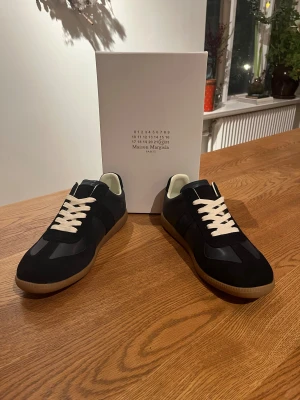 Margiela sneakers marinblåa - Margiela sneakers i marinblåmed kontrasterande vita snören och beige insida. Skorna har en klassisk rund tå och platt gummisula i brunt. Materialmix av skinn och mocka ger en lyxig känsla. Perfekt för dig som gillar stilrena och tidlösa sneakers.